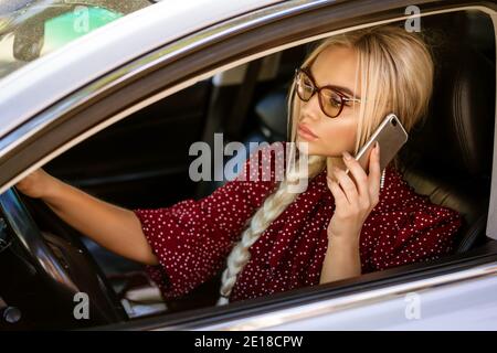 A blonde woman in glasses in a red blouse sits behind the wheel and solves business matters using the phone. Employment. Stock Photo