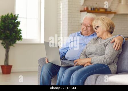 Loving happy elderly couple sitting together on sofa, hugging and watching movie online Stock Photo