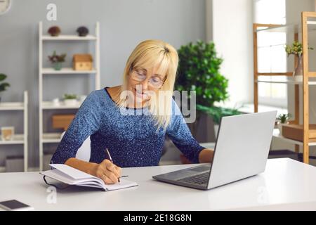 Senior woman making notes while looking at business training or online webinar on laptop. Stock Photo