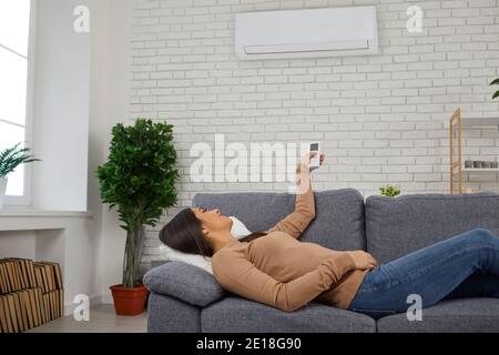 Woman lying on couch at home and switching on air conditioner with remote control Stock Photo