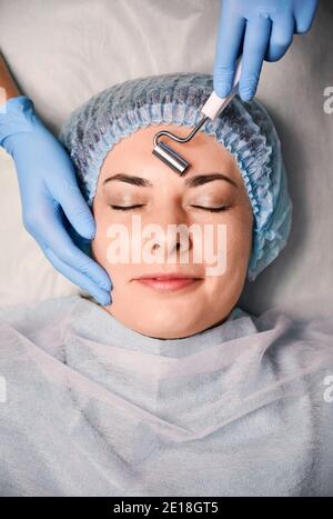 Close up of cosmetologist hands in sterile gloves using face roller massager or face lifting device while massaging female client face. Concept of skincare treatment, anti-aging procedure and beauty. Stock Photo