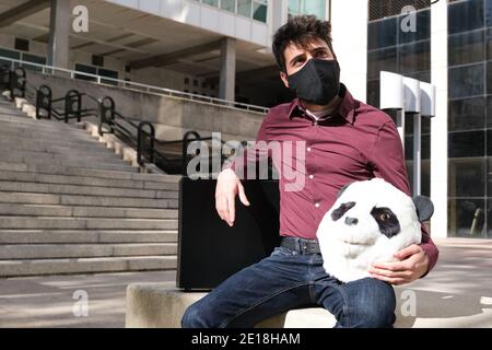 Young business man wearing face mask with a panda head mask in his hands and a suitcase, sitting on a bench. Bizarre businessman. Stock Photo