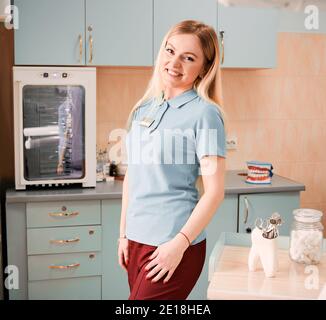 Beautiful young woman dentist looking at camera and smiling while posing in dental office, standing by the table with dental instruments. Concept of dentistry, stomatology and dental workers. Stock Photo