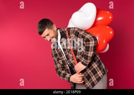 Young cute man carries a bunch of heart-shaped balloons on his back. St.Valentines Day on red background and side space. Stock Photo