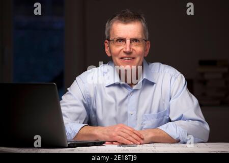 Smiling businessman or architect sitting by night at a desk with laptop and blueprint - focus on the face Stock Photo