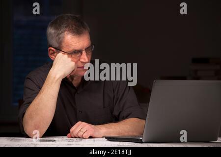 Mature businessman or engineer in dark shirt sitting by night at a desk looking at a laptop - dark image Stock Photo