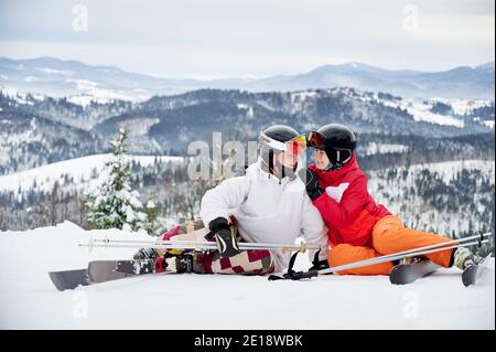Charming young woman looking at man and smiling while wrapping arm around his shoulder. Joyful boyfriend and girlfriend sitting on snow-covered hill in mountains. Concept of skiing and relationships.