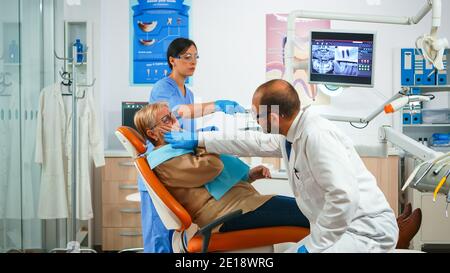 Old patient touching cheek showing tooth with pain, complaining about aching tooth. Elderly woman explaining dental problem to doctor indicating mouth while sitting on stomatological chair Stock Photo