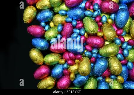 Pile or group of multi coloured & different sizes of colourful foil wrapped chocolate easter eggs in pink, blue, yellow and lime green against black. Stock Photo