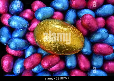 Pile or group of multi colored and different sizes of colourful foil wrapped chocolate easter eggs in pink, and blue, with a large yellow gold egg in Stock Photo