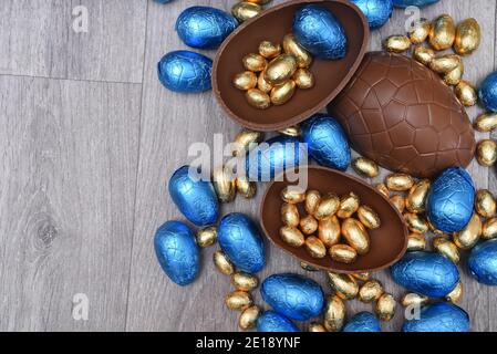 Pile or group of different sizes of colourful foil wrapped chocolate easter eggs in blue, and gold. Large halves of a brown milk chocolate egg have mi Stock Photo
