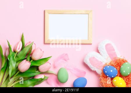 Easter eggs in a nest, bunny ears, tulips and a frame with copy space on a pink background. Flat lay greeting card. Stock Photo