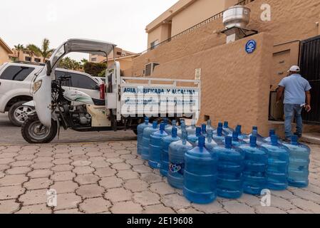 A man dressed in blue jeans and blue shirt delivers bottles of purified water in his trike cargo truck, large blue bottles lined up on the pavement. Stock Photo