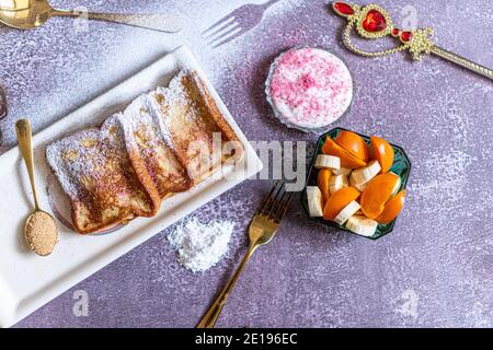 Traditional french toast with cinnamon on a gray background. Traditional Spanish sweet fried toasts, torrijas, with banana and persimmon, fresh cream, powdered sugar on a ceramic plate, and gold cutlery. Typical dessert for Lent and Easter. Top view. High quality photo  Stock Photo