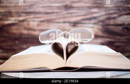 Old opened hardback book  with heart form sheets and reading glasses, isolated on a wooden background. Love reading. Close up Heart from book Stock Photo