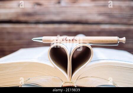 Old opened hardback book with heart form sheets, reading glasses and a pen isolated on a wooden background. Love reading. Close up Heart from book Stock Photo