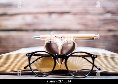 Old opened hardback book with heart form sheets, reading glasses and a pen isolated on a wooden background. Love reading. Close up Heart from book Stock Photo