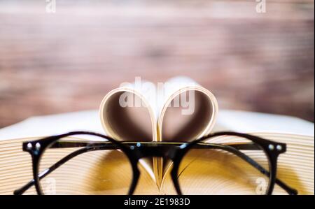 Old opened hardback book  with heart form sheets and reading glasses, isolated on a wooden background. Love reading. Close up Heart from book Stock Photo