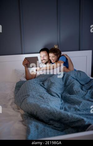 Beautiful lovely playful young couple having fun with a tablet in bed at night. Relationship and weekend goals. Stock Photo