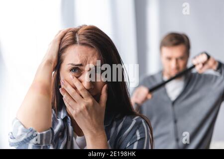 Worried woman with bruise on hand covering mouth near abusive husband with waist belt on blurred background
