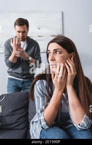 Scared victim of domestic violence with bruise on cheek sitting on couch near husband on blurred background Stock Photo