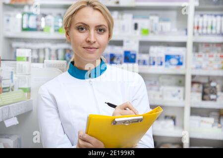 Confident female pharmacist taking notes on her clipboard, smiling to the camera joyfully. Lovely female chemist working at her drugstore, checking st Stock Photo