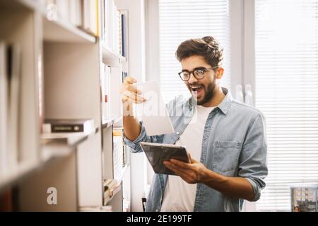 Handsome nerdy young cheerful student in library celebrating for getting the newest book. Stock Photo