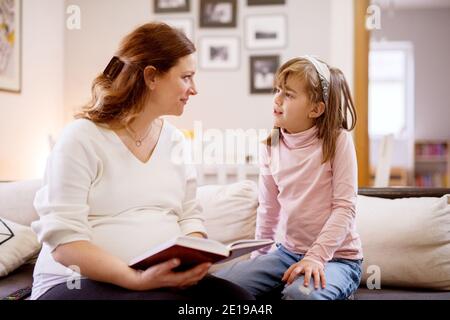 Sweet cheerful little toddler girl sitting on the sofa and looking curious while her beautiful pregnant mother reading a story. Family and new life co Stock Photo