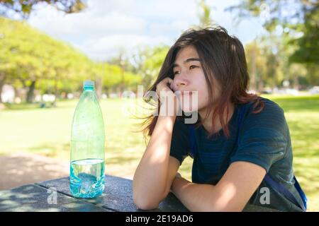 Biracial Asian Caucasian teen girl sitting in shade outdoors under trees on sunny day, resting chin on hand at picnic table, thinking Stock Photo
