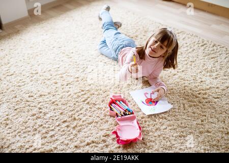 Adorable pretty little toddler girl drawing on paper with wooden crayons while lying on the carpet in her room. Stock Photo