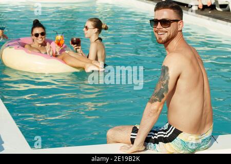 Handsome tattooed young man smiling to the camera over his shoulder, sitting on the edge of a swimming pool, his female friends having fun in the wate Stock Photo