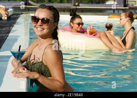 Gorgeous tanned young woman smiling to the camera joyfully, having a cocktail at the pool party, her friend swimming on inflatable donut on the backgr Stock Photo