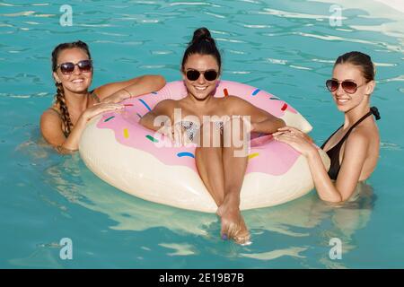 Three happy beautiful female friends having a pool party, smiling to the camera, swimming on a inflatable donut together. Gorgeous women enjoying day Stock Photo