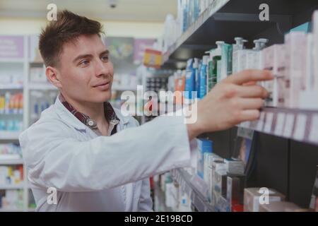Cheerful handsome male pharmacist doing stocktaking, organizing products on shelves at drugstore. Charming young male chemist working at the pharmacy. Stock Photo