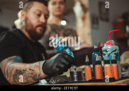 Selective focus on tattoo ink in bottles, tattooist working at his studio, copy space on the side. Young man getting new tattoo on chest. Professional Stock Photo