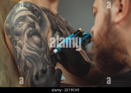 Cropped close up of a bearded tattoo artist working at his studio tattooing sleeve on the arm of his male client. Man getting tattooed by professional Stock Photo