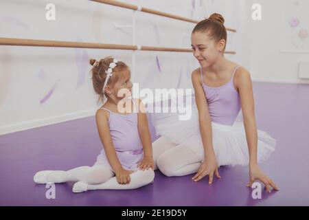 Two lovely young ballerinas smiling at each other, resting on the floor after dancing at ballet school. Adorable little ballerina and her sister weari Stock Photo
