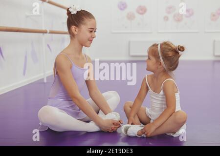 Two adorable little ballerinas smiling at each other, sitting on the floor, resting after dancing. Cute little ballerina girl smiling at her older sis Stock Photo