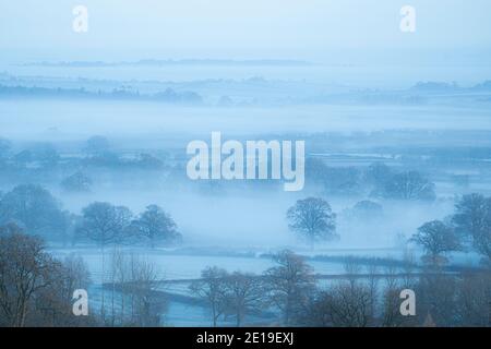 Misty blue landscape view of English countryside with mist layers rolling over trees and fields, rural scene of beautiful mist layered beautiful peaceful British scenery in The Cotswolds, England, UK Stock Photo