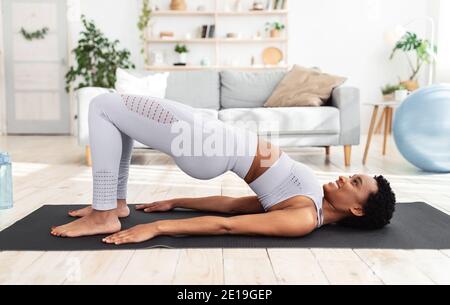 Black woman doing pilates workout using elastic strap sitting on yoga mat,  pulling training arms and shoulders in home living room. Athletic fit  exercising body using resistance band. Stock Photo