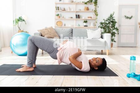 Black expectant lady doing abs exercises on mat at home, standing in half bridge pose, taking care of her future child Stock Photo