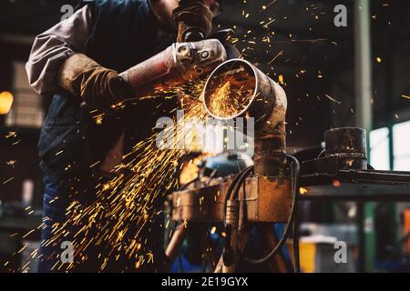 Fabric worker in protective uniform cutting metal pipe on the work table with an electric grinder in the industrial workshop. Stock Photo