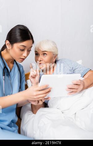excited senior woman showing idea gesture while using digital tablet together with astonished asian nurse Stock Photo