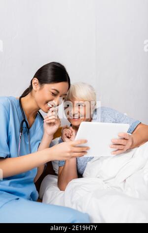 excited asian nurse and senior woman laughing while using digital tablet together Stock Photo