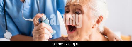 elderly woman with open mouth suffering from asthma attack and using inhaler near nurse on blurred background, banner Stock Photo