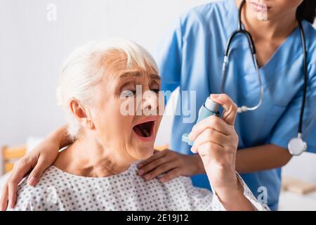 elderly woman using inhaler while suffering from asthma attack near nurse touching her shoulders on blurred background Stock Photo