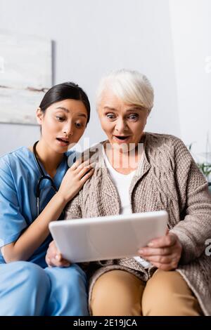 surprised asian woman touching shoulder of amazed senior woman looking at digital tablet Stock Photo