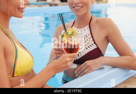 Cropped shot of two happy beautiful women enjoying cocktails at the swimming pool. Cheerful female friends relaxing at the poolside, having drinks tog Stock Photo