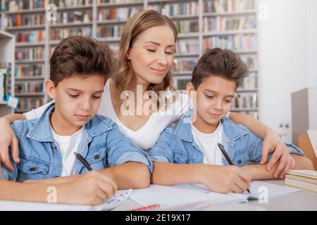 Beautiful happy woman hugging her two twin sons, while they write in their textbooks at the library Stock Photo