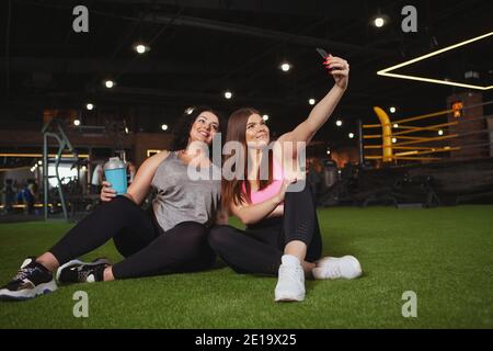 Lovely happy plus size sportswomen resting at the gym after exercising, taking selfies on smart phone. Charming curvy ladies relaxing at sports studio Stock Photo